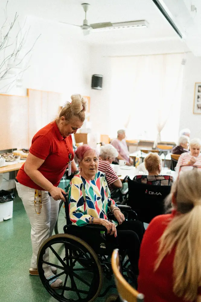 A caregiver assists an elderly woman in a wheelchair in a communal setting filled with other seniors.