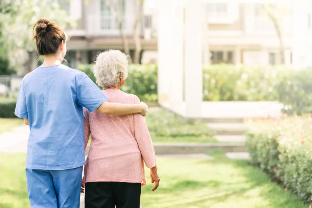 A caregiver gently assists an elderly woman as they walk together in a sunlit garden.