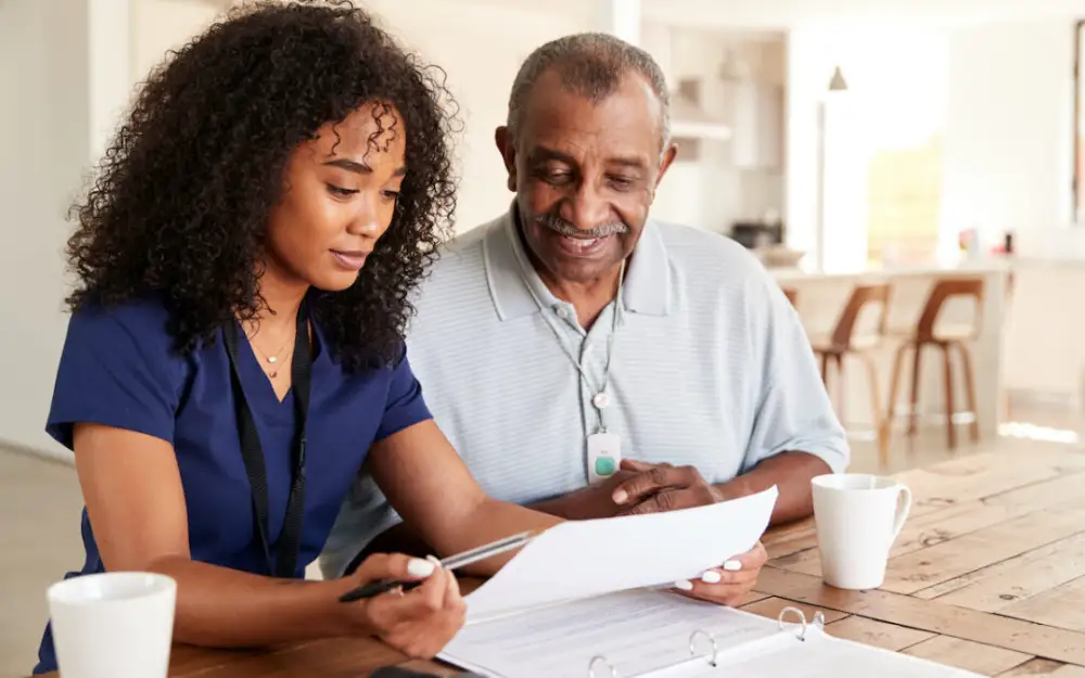 A woman in scrubs assists an older man with paperwork at a table, fostering a supportive conversation.