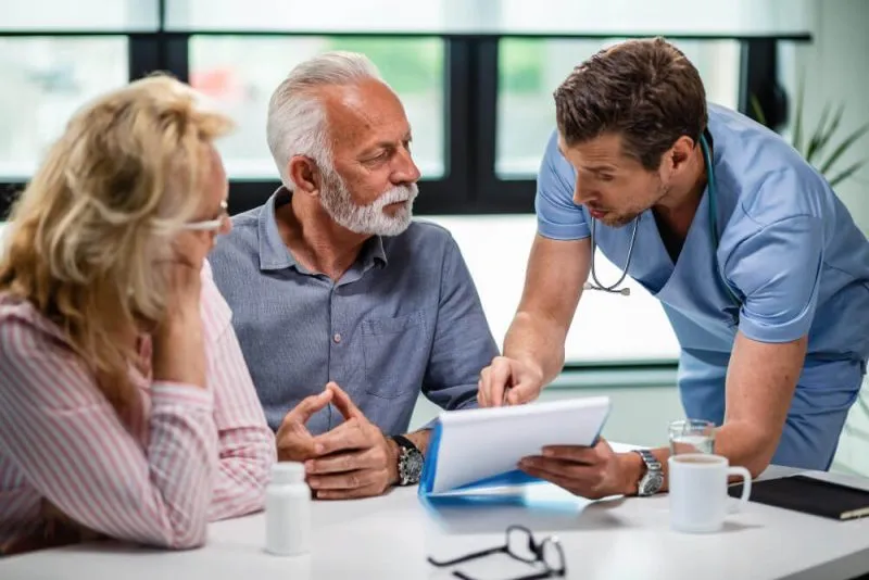 A doctor explains medical information to an elderly couple during a consultation in a bright office setting.