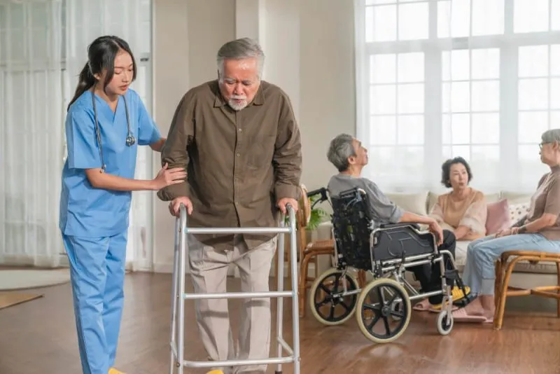 A caregiver assists an elderly man with a walker in a bright room with other seniors seated nearby.