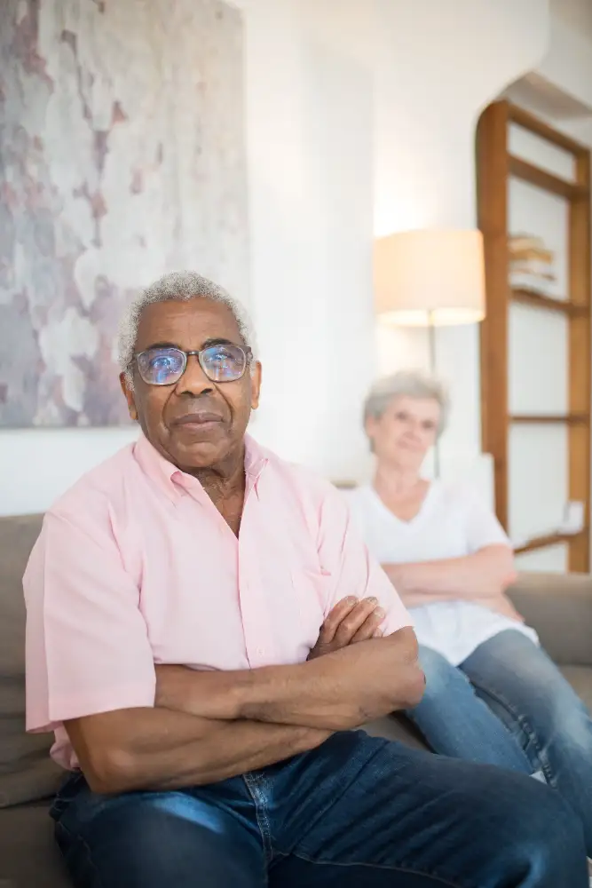 An older man in a pink shirt sits with crossed arms, while a woman in the background appears disengaged.
